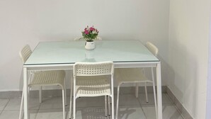 Een eethoek met een glazen tafel, witte stoelen, een vaas met bloemen, een klok en een ingelijst schilderij aan de muur.