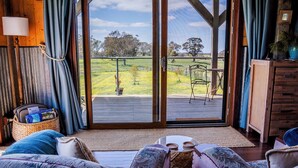 A room with a large sliding glass door leading to an outdoor deck with a metal chair. There is a wicker basket with items on the floor, a wooden cabinet, and a bed with patterned bedding. - Stunningly built open-plan Studio in natural pastureland setting (Hay Valley)