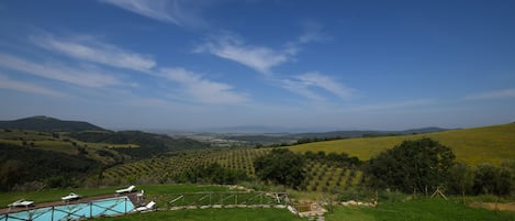 A pool area with a stone pathway, surrounded by lush green fields and distant hills under a clear sky.