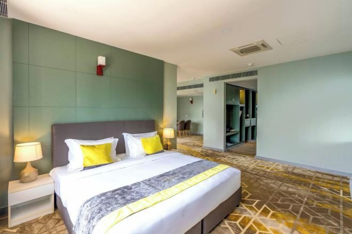 Habitación de hotel con una cama grande, una mesita de noche con lámpara y vistas al pasillo y otras habitaciones.