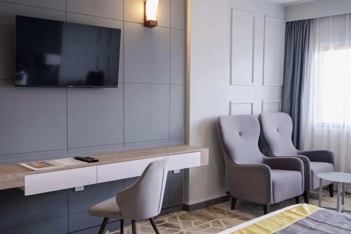 Una habitación de hotel moderna con una televisión de pantalla plana montada en la pared, un escritorio de madera con una silla, dos sillones grises y una mesita redonda.