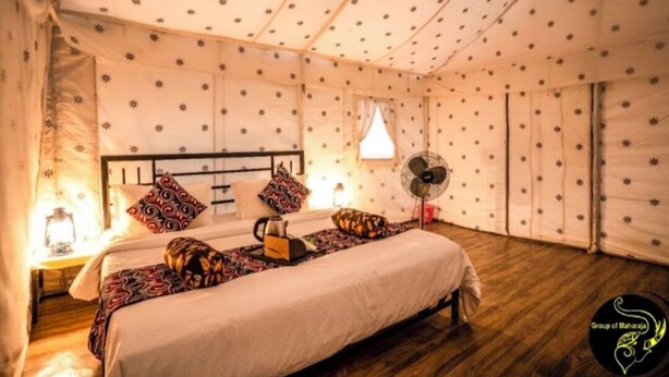 A cozy room with a bed, pillows, a bedside table with a lamp, and a fan. - Star Desert Camp Jaisalmer  (Jaisalmer)
