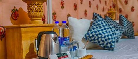A hotel room with a wooden nightstand, a kettle, two bottles of water, a towel, and a bed with blue and white checkered pillows.