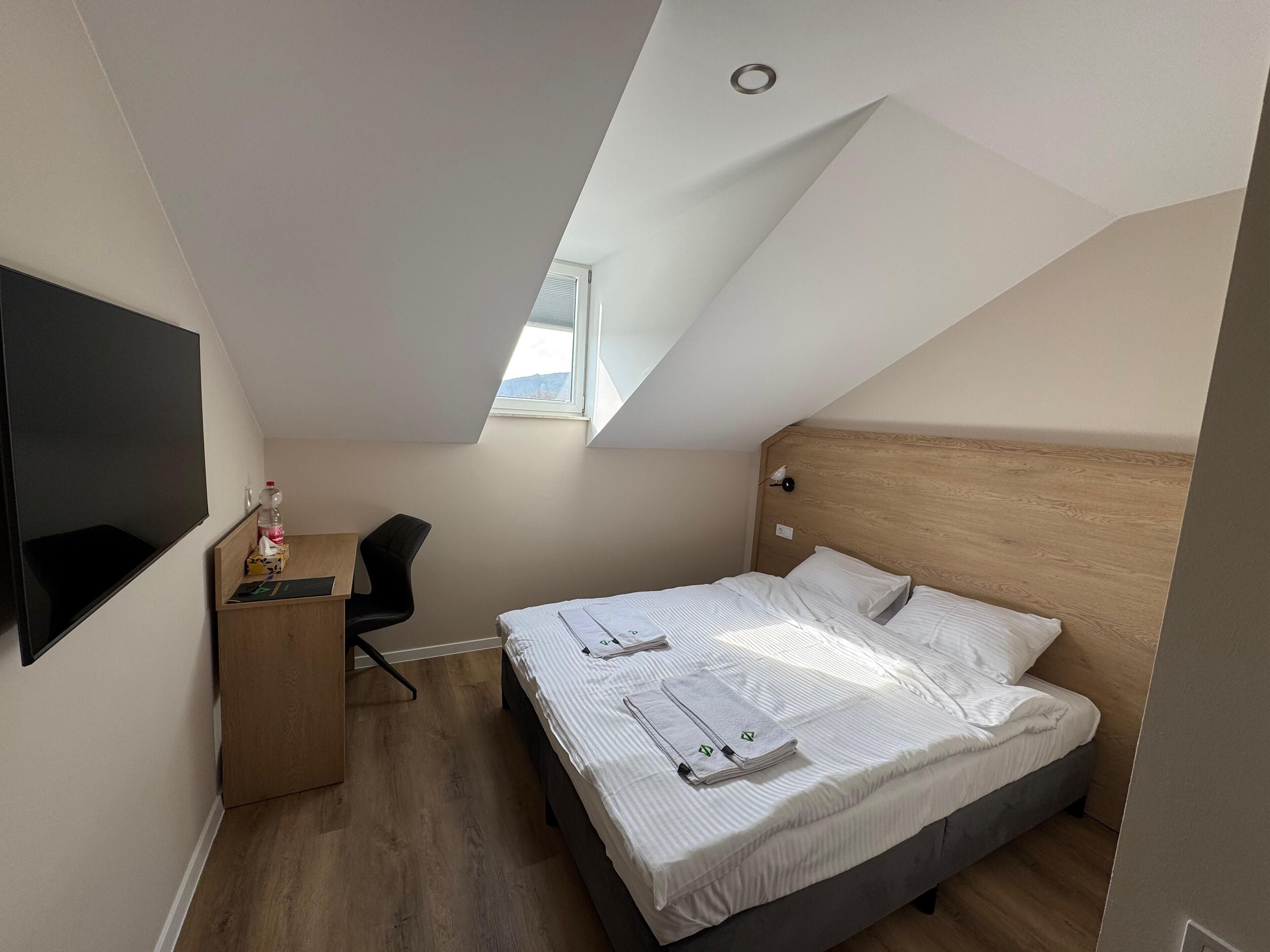 A bedroom with a sloped ceiling, a bed with white linens, a desk with a chair, and a TV mounted on the wall.