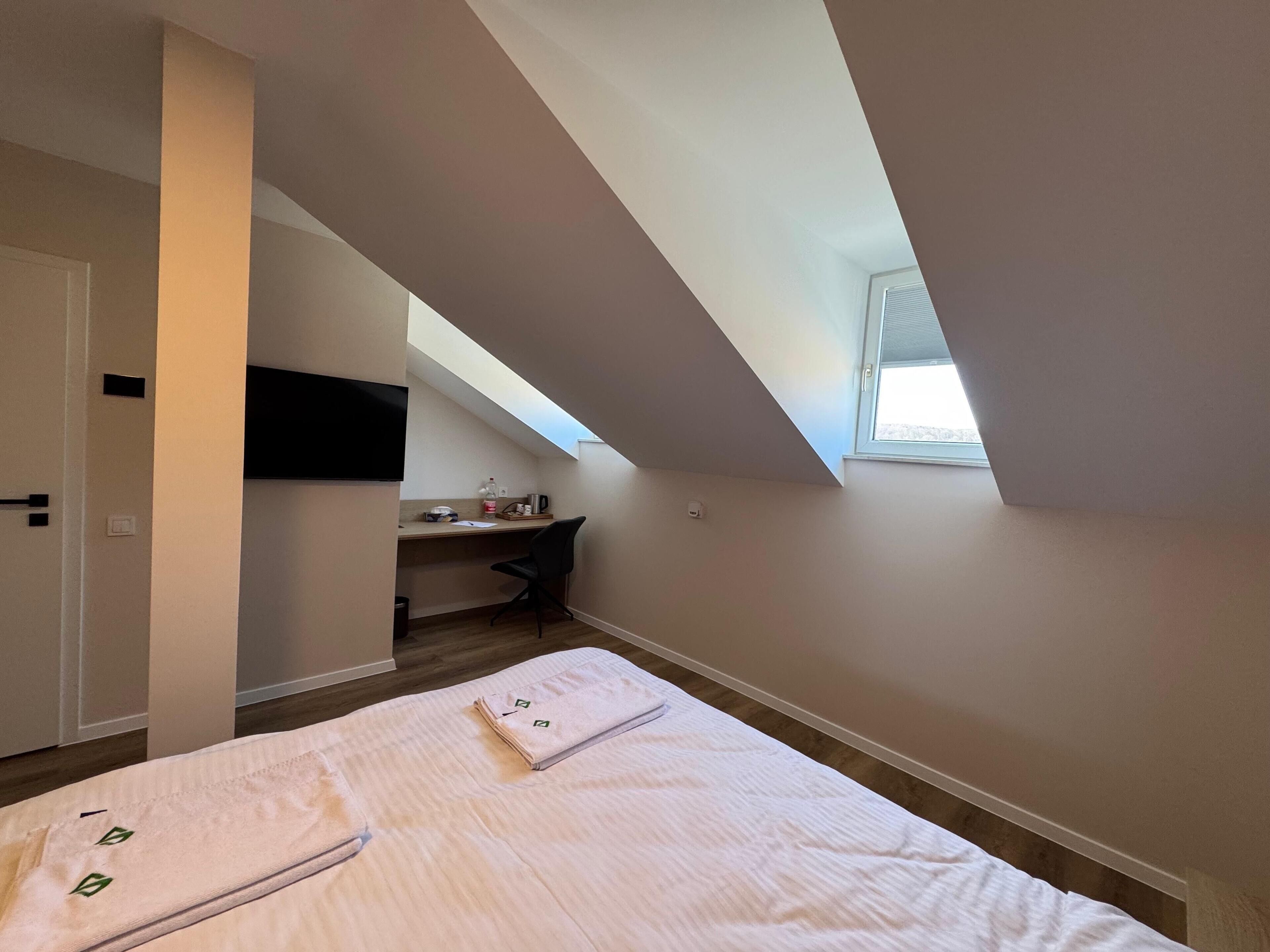 A bedroom with a sloped ceiling, a bed with a white bedspread, a desk with a chair, and a window providing natural light.