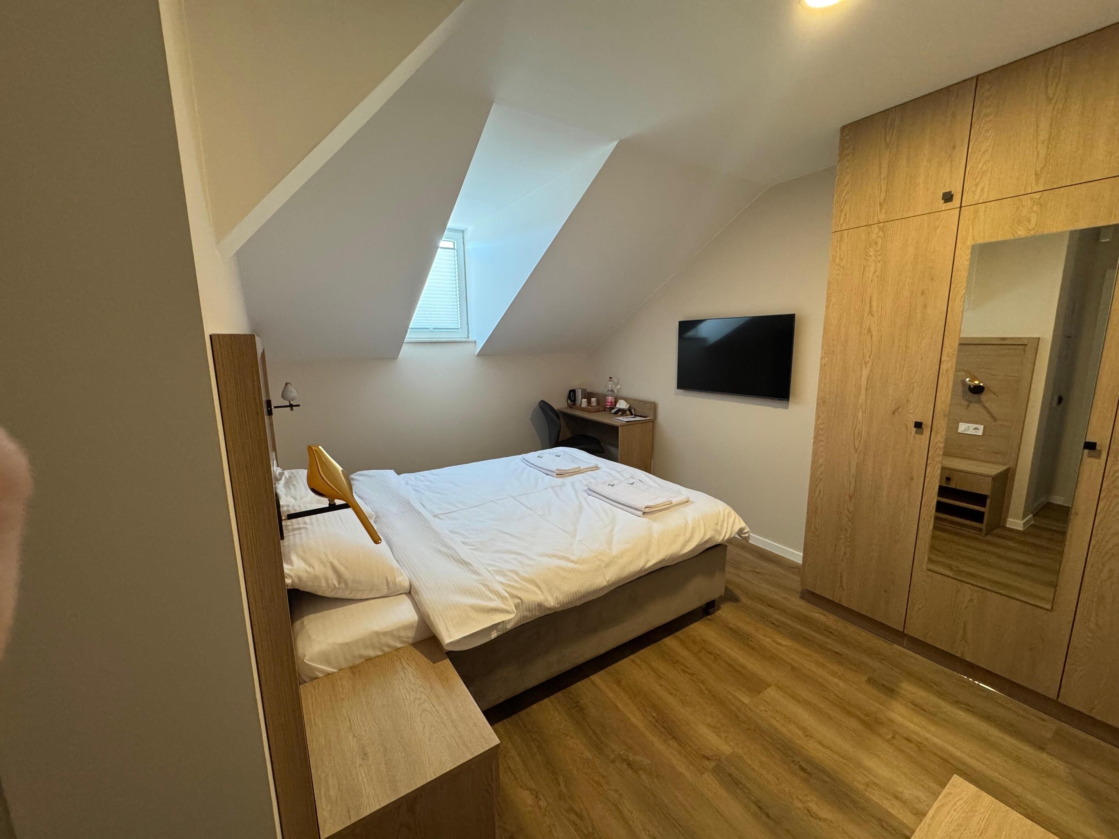 Ett sovrum med ett sluttande tak, en säng, ett skrivbord, en stol, en tv och en garderob i trä.