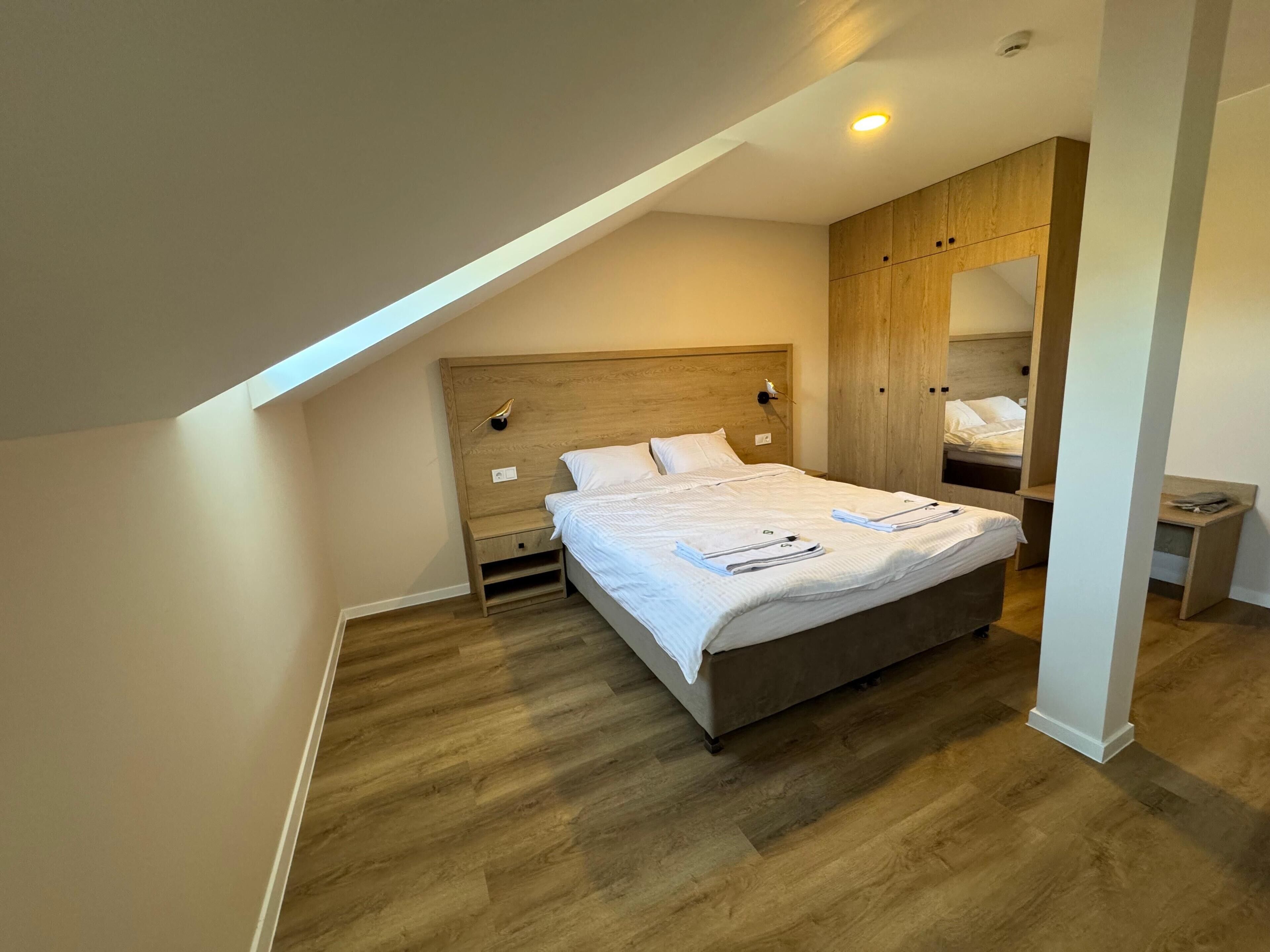 Quarto moderno com teto inclinado, cabeceira de madeira e cama com roupa de cama bem dobrada.