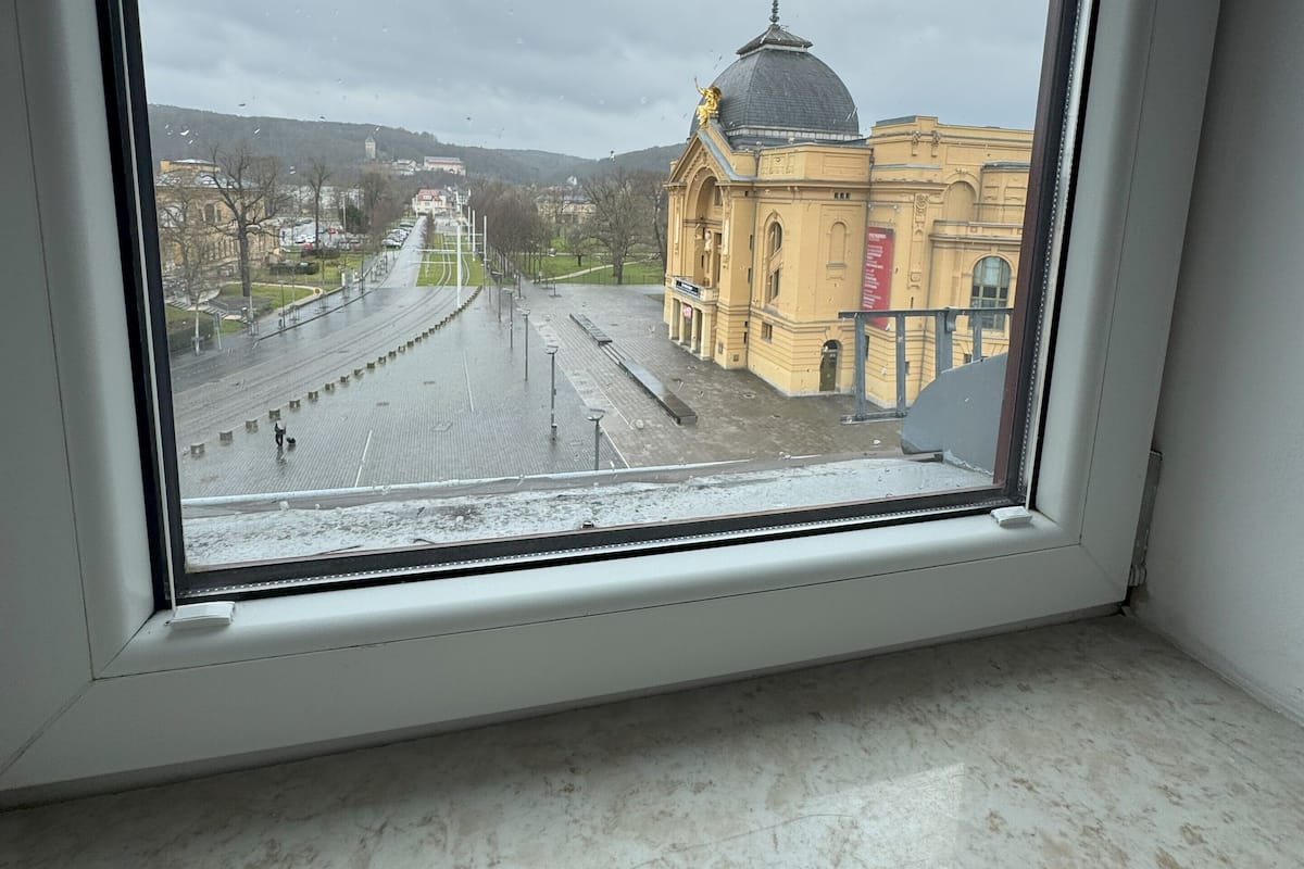 A view through a window of a building with a dome and a statue on top, a paved area with a fence, and a cloudy sky.
