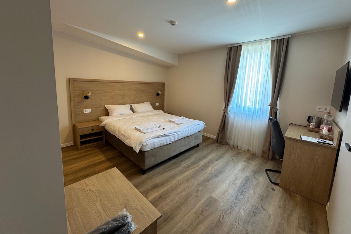 A hotel room with a wooden bed, bedside tables, a desk, a TV, and a window with curtains.