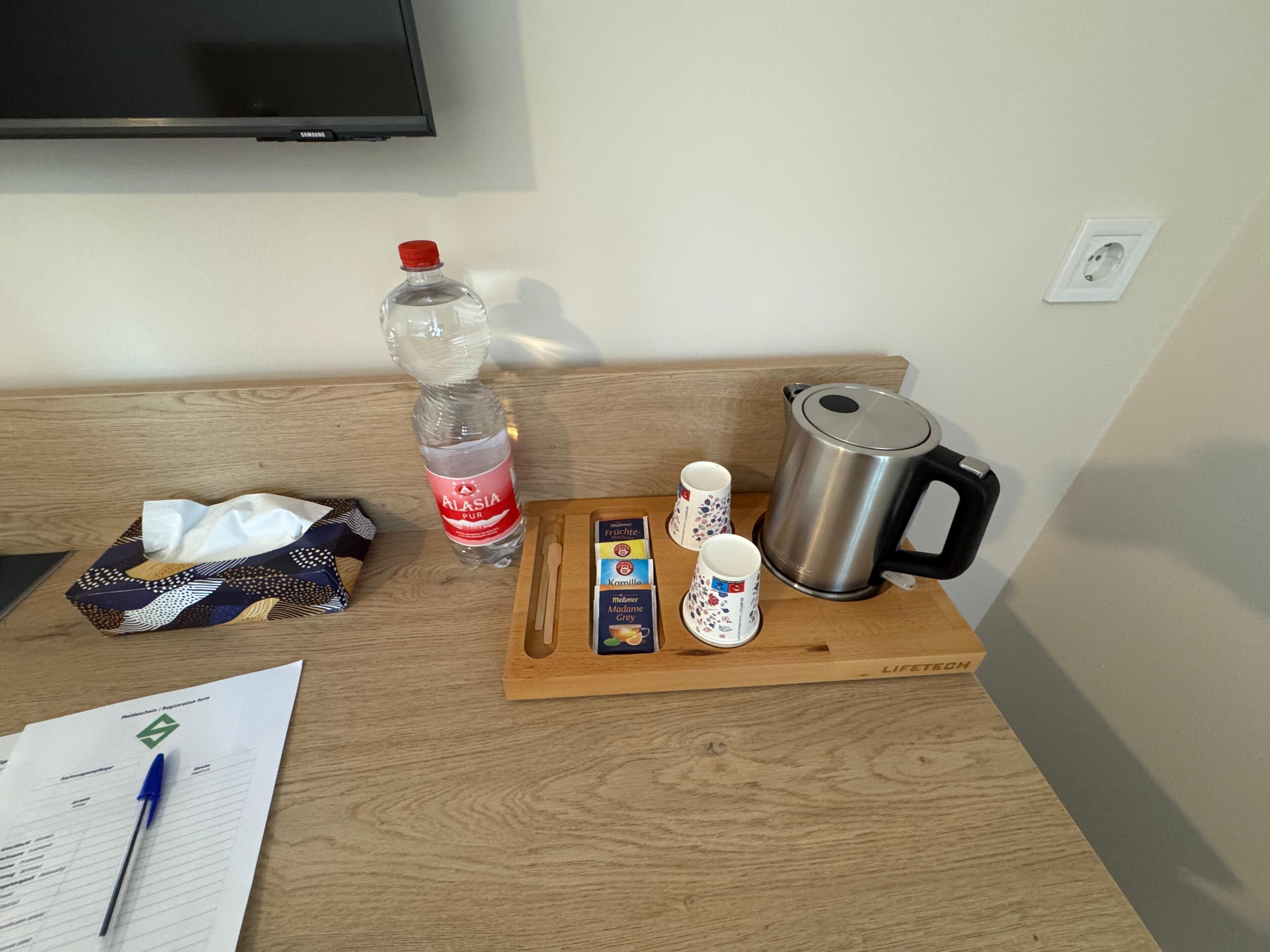 A wooden table with a bottle of water, a kettle, a tray with cups and a box of tea, and a registration form with a pen.