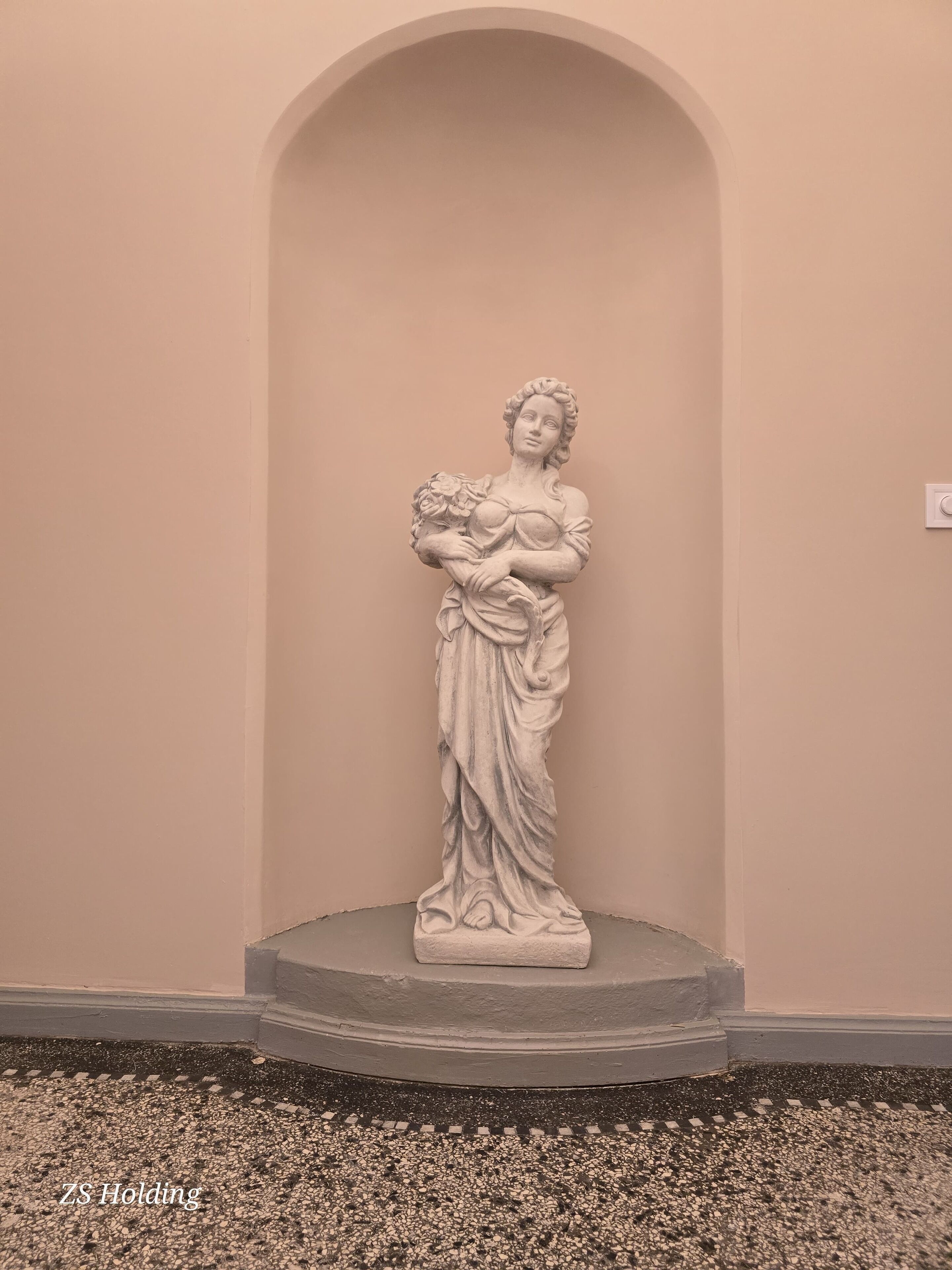 Eine Statue einer Frau, die ein Kind hält, auf einem Sockel in einem Raum mit schlichter Wand und Fliesenboden.