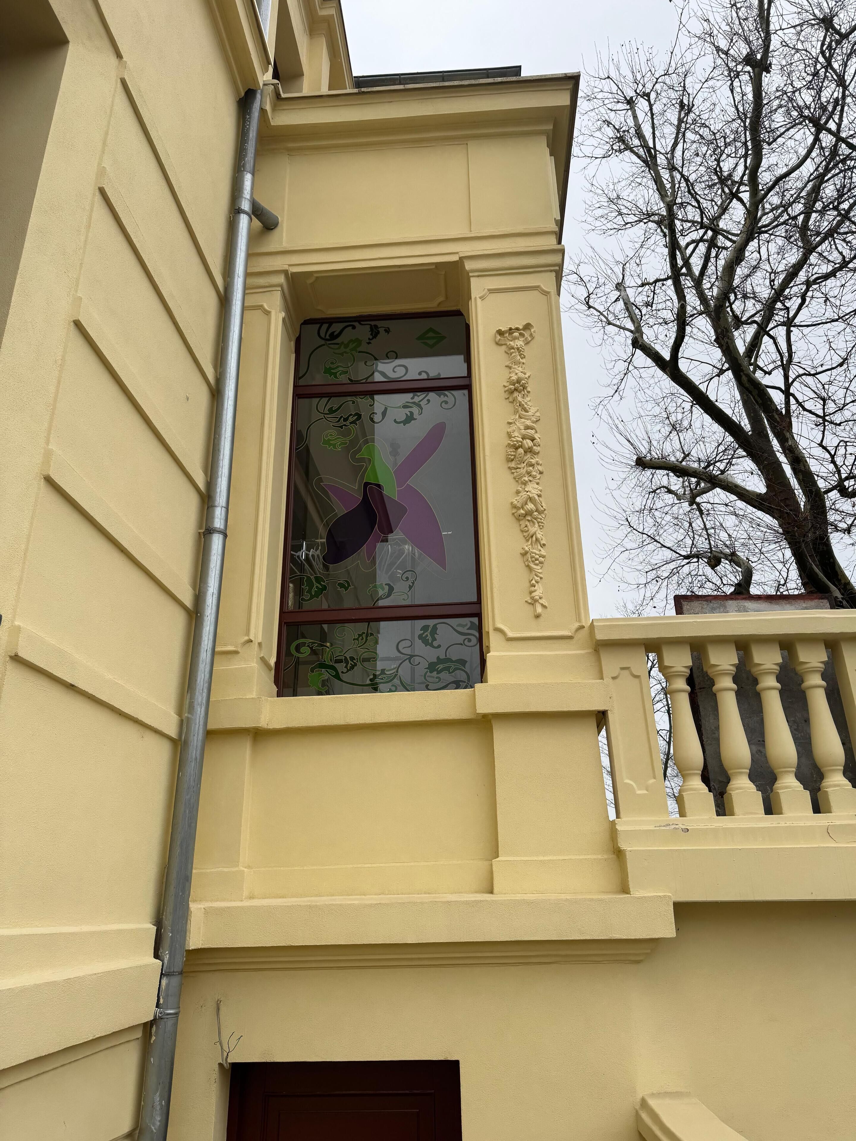 Un bâtiment à la façade jaune, une fenêtre ornée de vitraux avec un motif en éventail violet, et un arbre en arrière-plan.