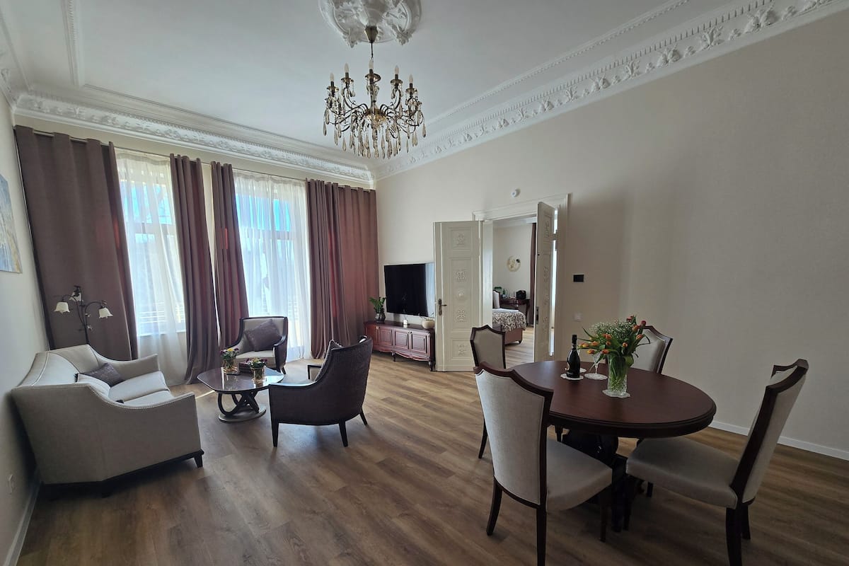 A spacious living room with a chandelier, a dining table with chairs, and a television in the background.