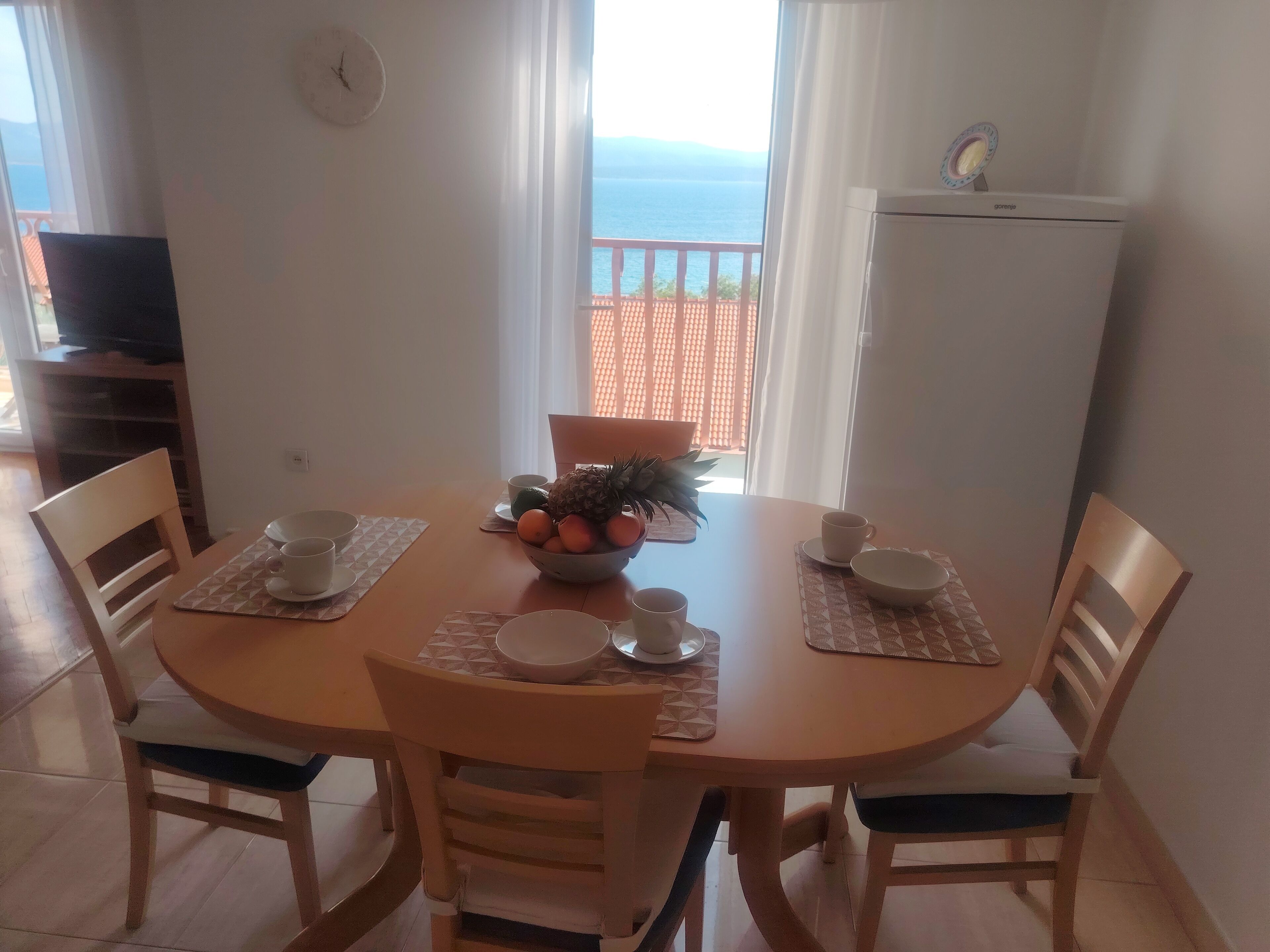 A dining table with a bowl of fruit, cups, and a clock on the wall.