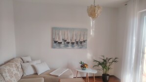 Un salon comprenant un canapé, une table basse, une petite table d’appoint avec un saladier de fruits, une plante en pot et un tableau représentant des voiliers accroché au mur.
