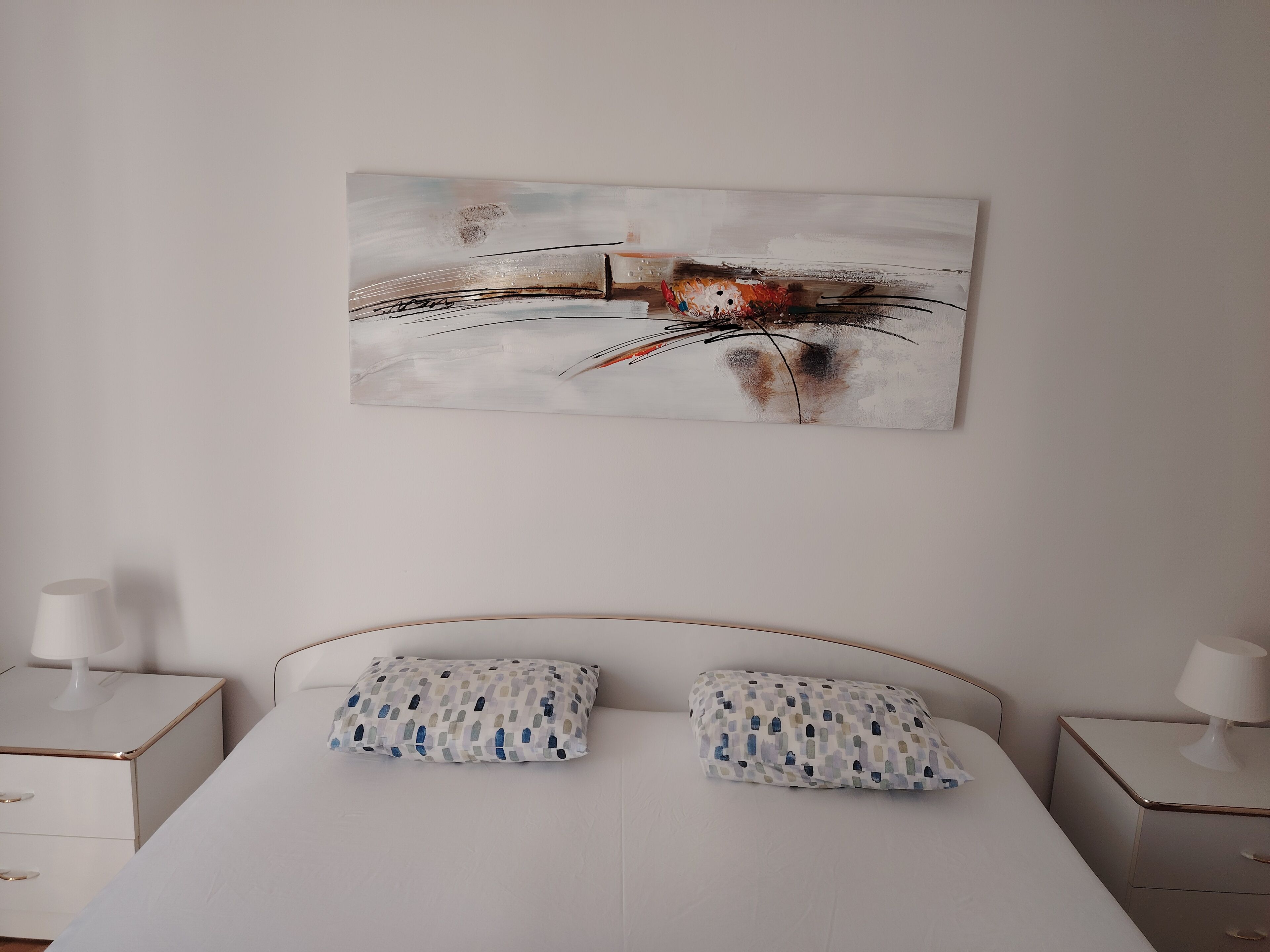 A bedroom with a bed, two pillows, a nightstand, and a wall-mounted abstract painting.