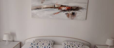 A bedroom with a bed, two pillows, a nightstand, and a wall-mounted abstract painting.