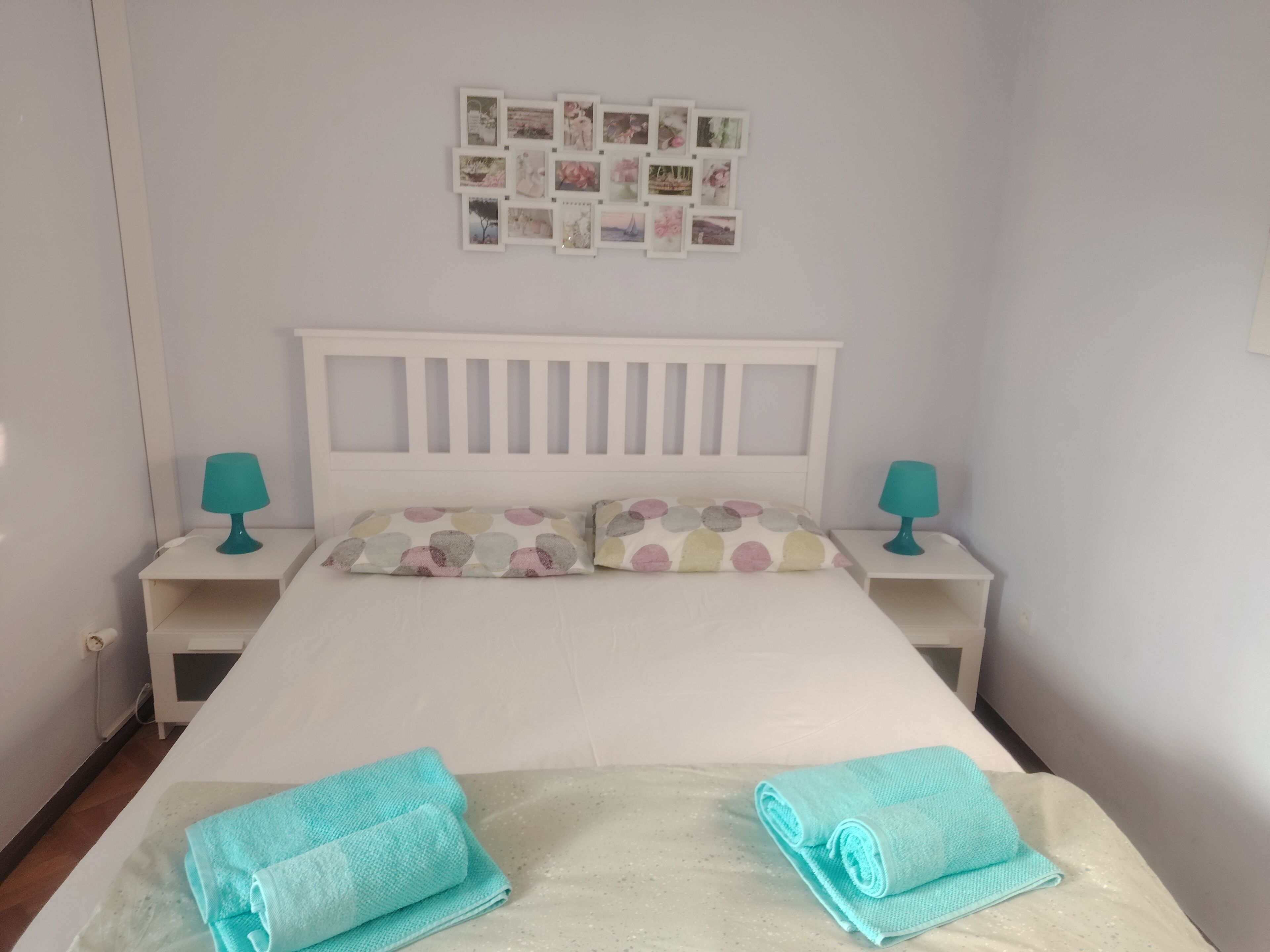 A neatly made bed with a white headboard, two matching teal lamps on nightstands, and a wall of framed photos.