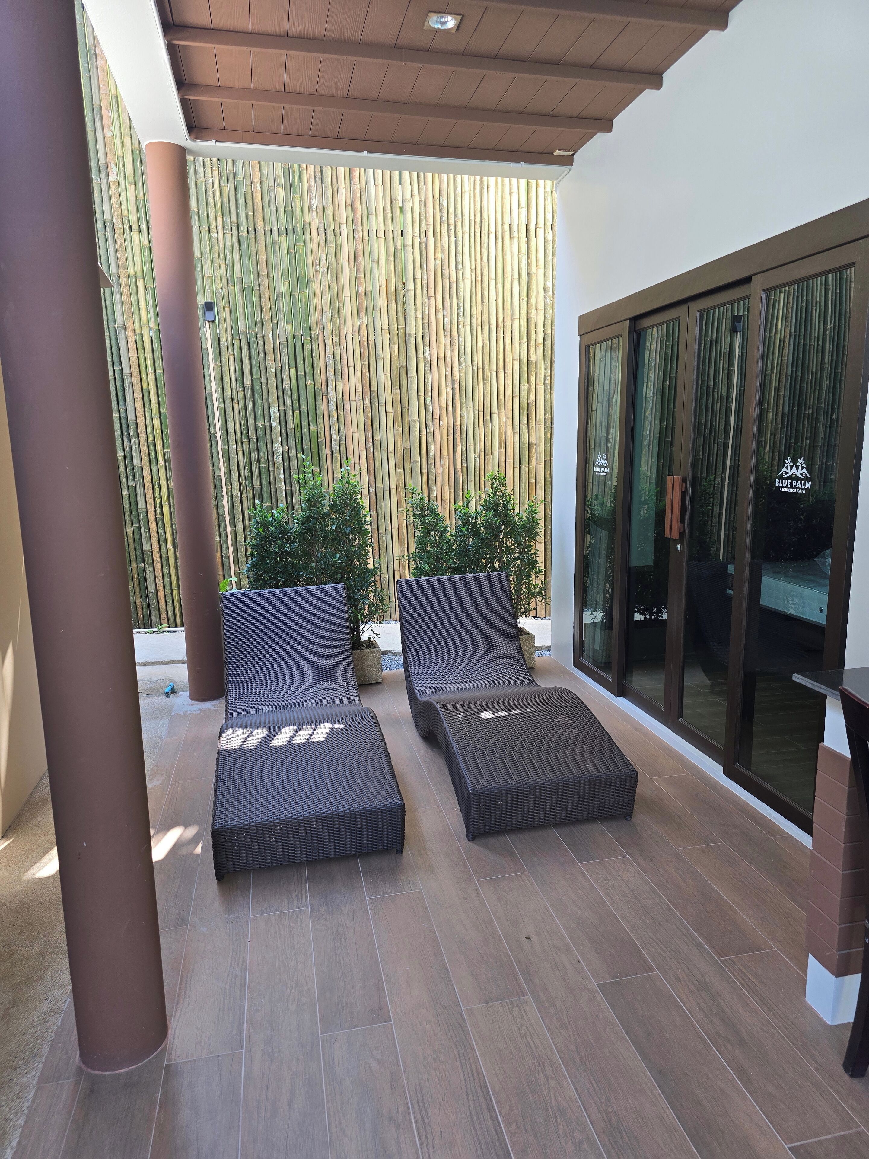 A covered patio area with two dark wicker lounge chairs, a bamboo wall, and a glass door leading to a room labeled "Blue Palm Residences".