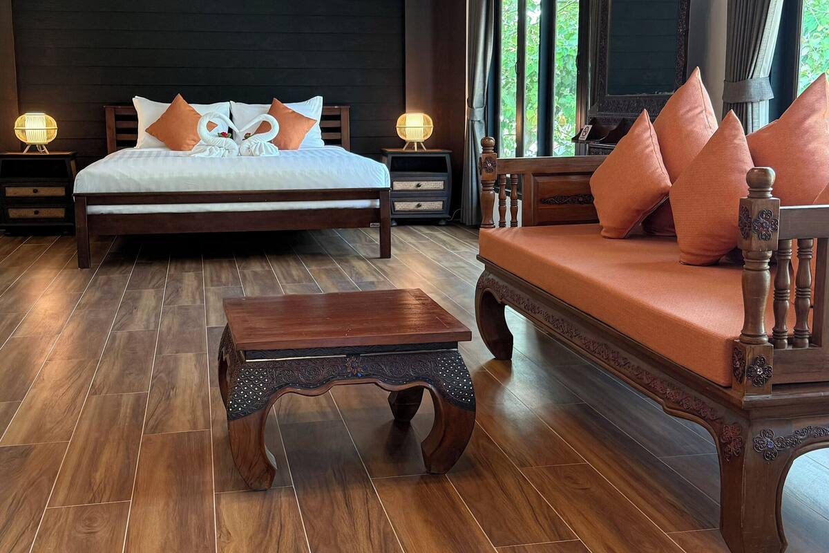 A spacious room with a large bed, a wooden sofa, and a small table. There is a ceiling fan, a window with curtains, and a wall-mounted air conditioner.