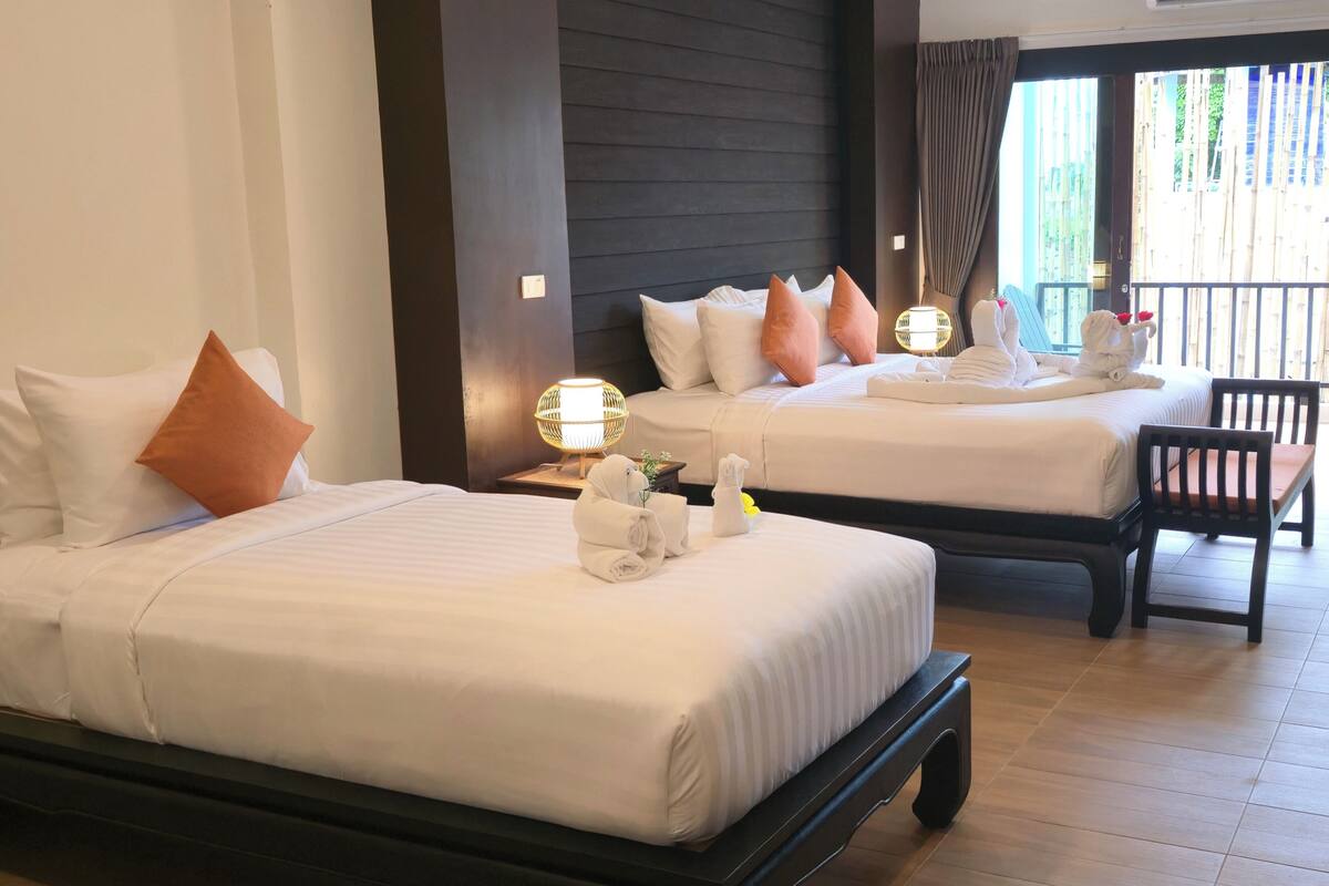A hotel room with two beds, each with white linens and a single orange pillow. There is a wooden nightstand with a lamp between the beds.