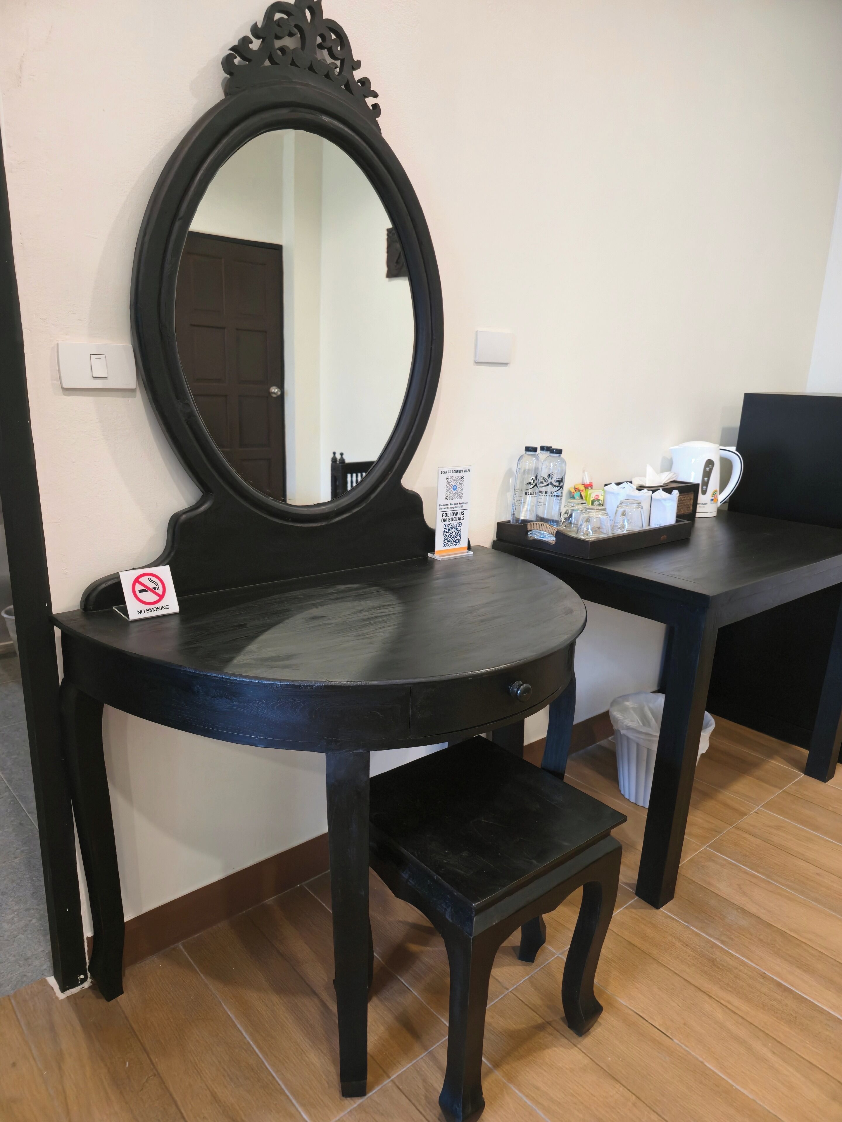 A room with a round table, a mirror, a "No Smoking" sign, a trash bin, and a coffee station with cups, bottles, and a kettle.