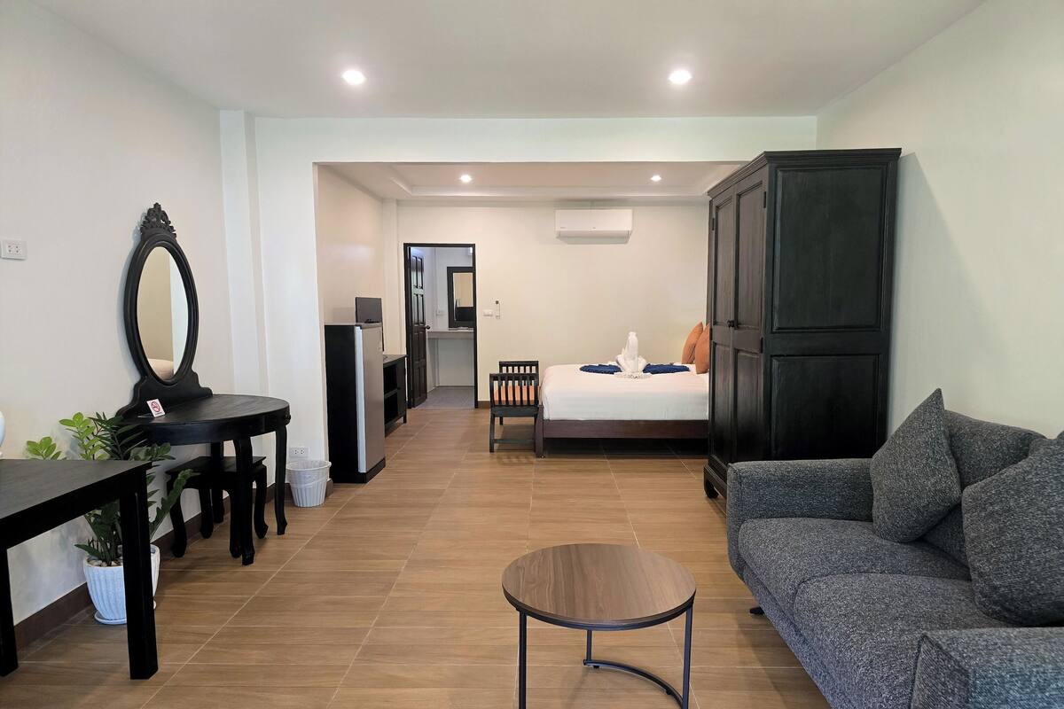 A modern living room with a sofa, a round coffee table, a dining table with chairs, a mirror, a refrigerator, and a bed with pillows.
