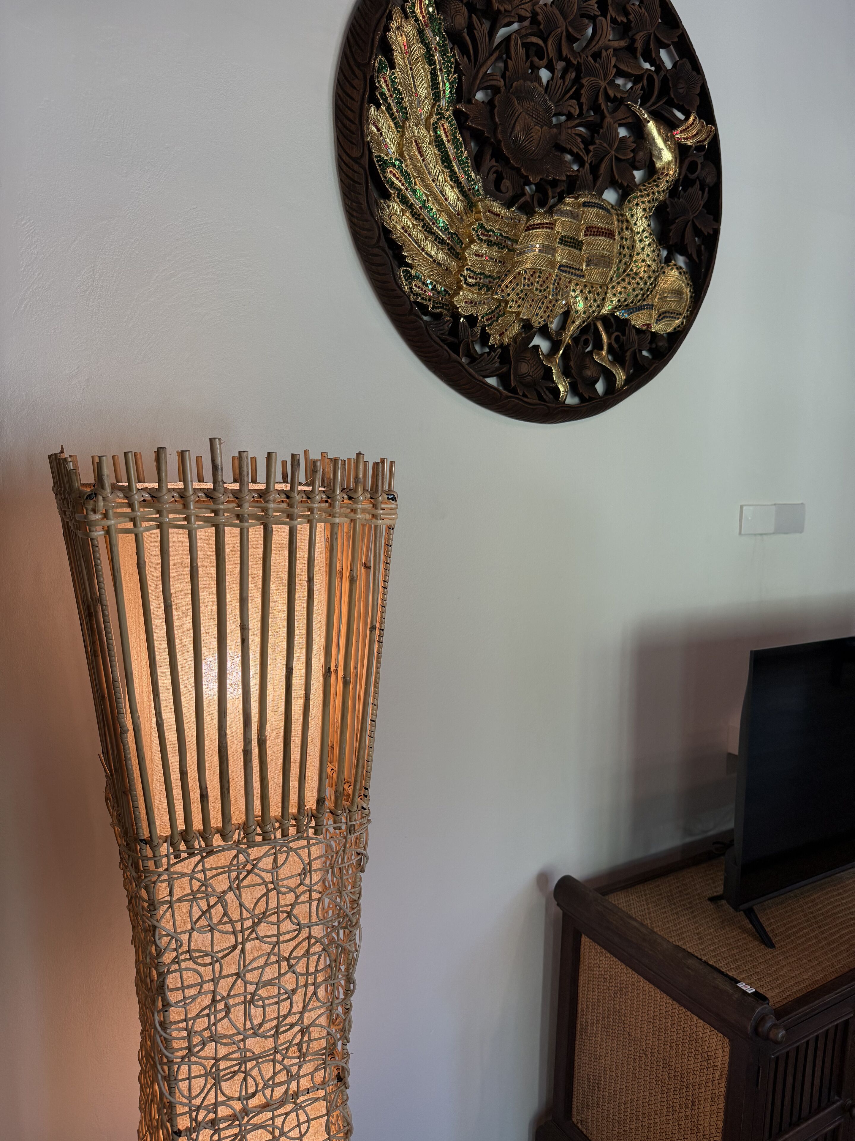 A room with a tall, woven lamp, a decorative wall piece, and a wooden cabinet with a screen.