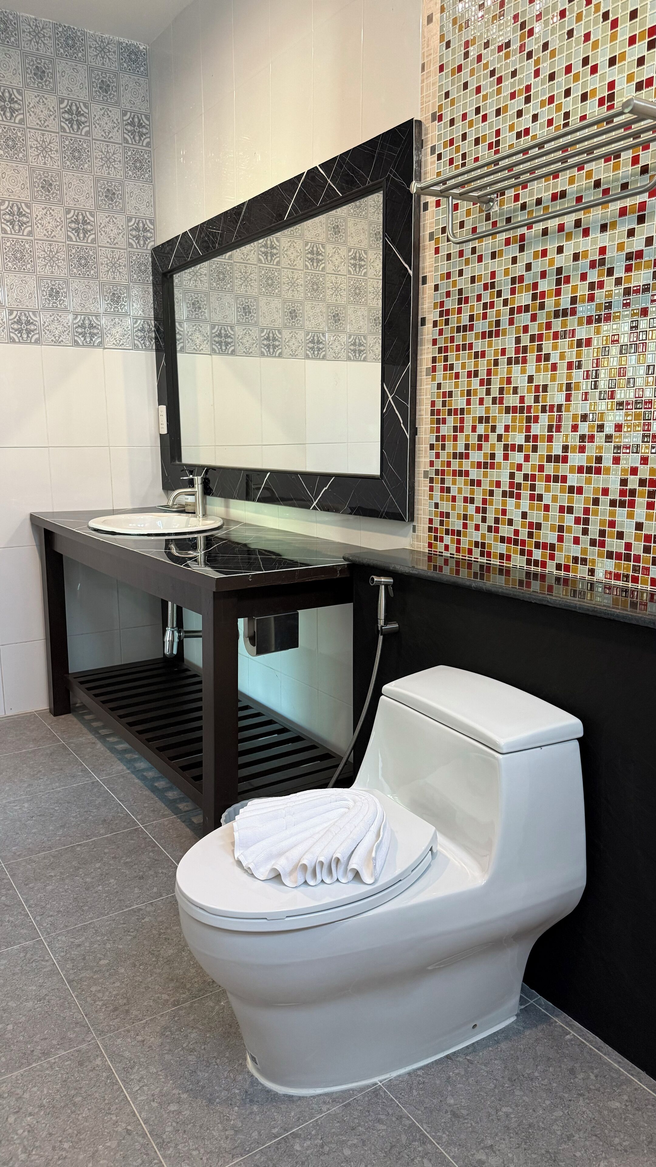 Un baño moderno con inodoro blanco, tocador negro con lavabo de vidrio y salpicadero de azulejos con diseño tipo mosaico.