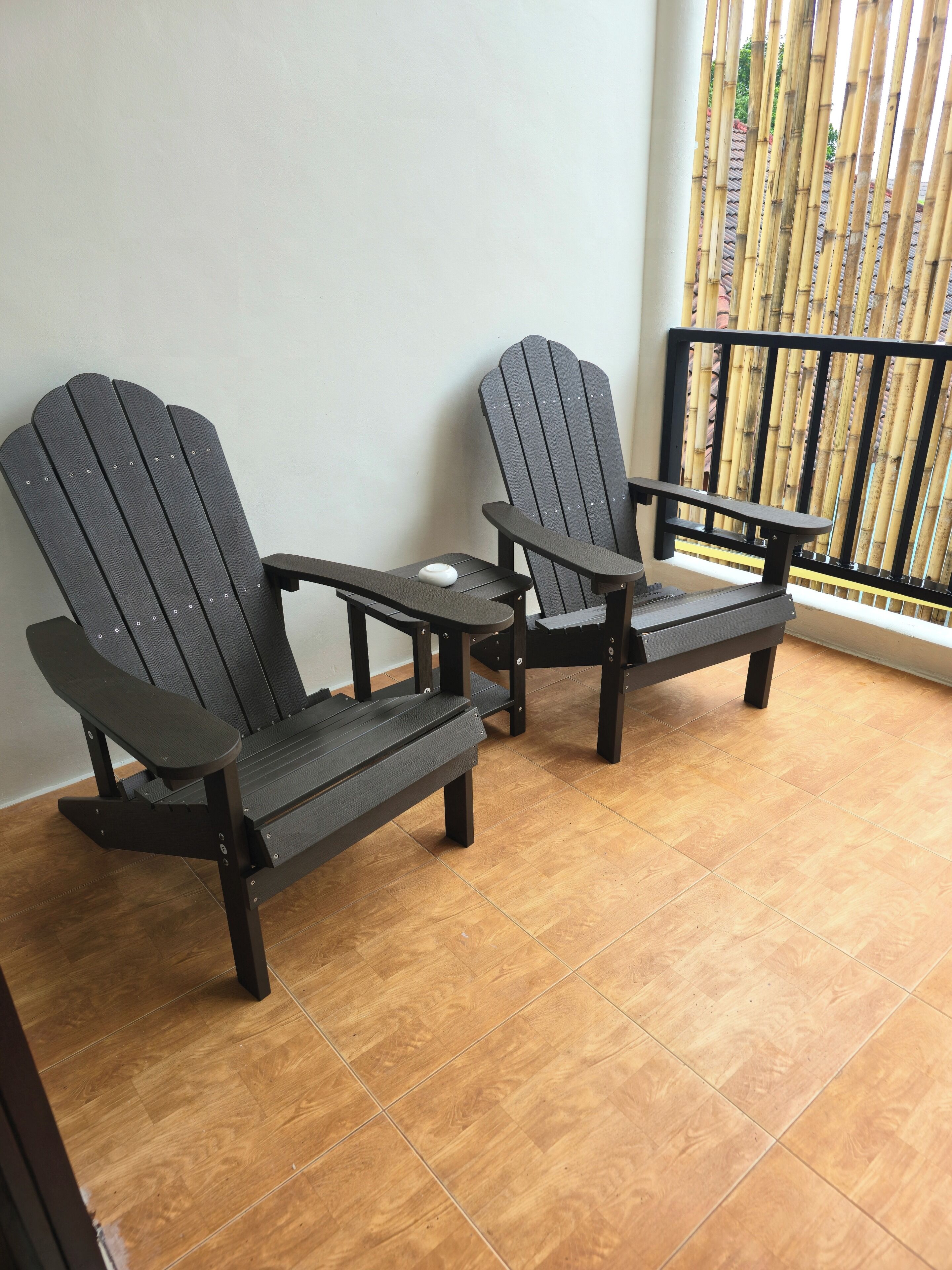 Two Adirondack chairs with a small table in between, positioned on a tiled balcony with a bamboo fence in the background.