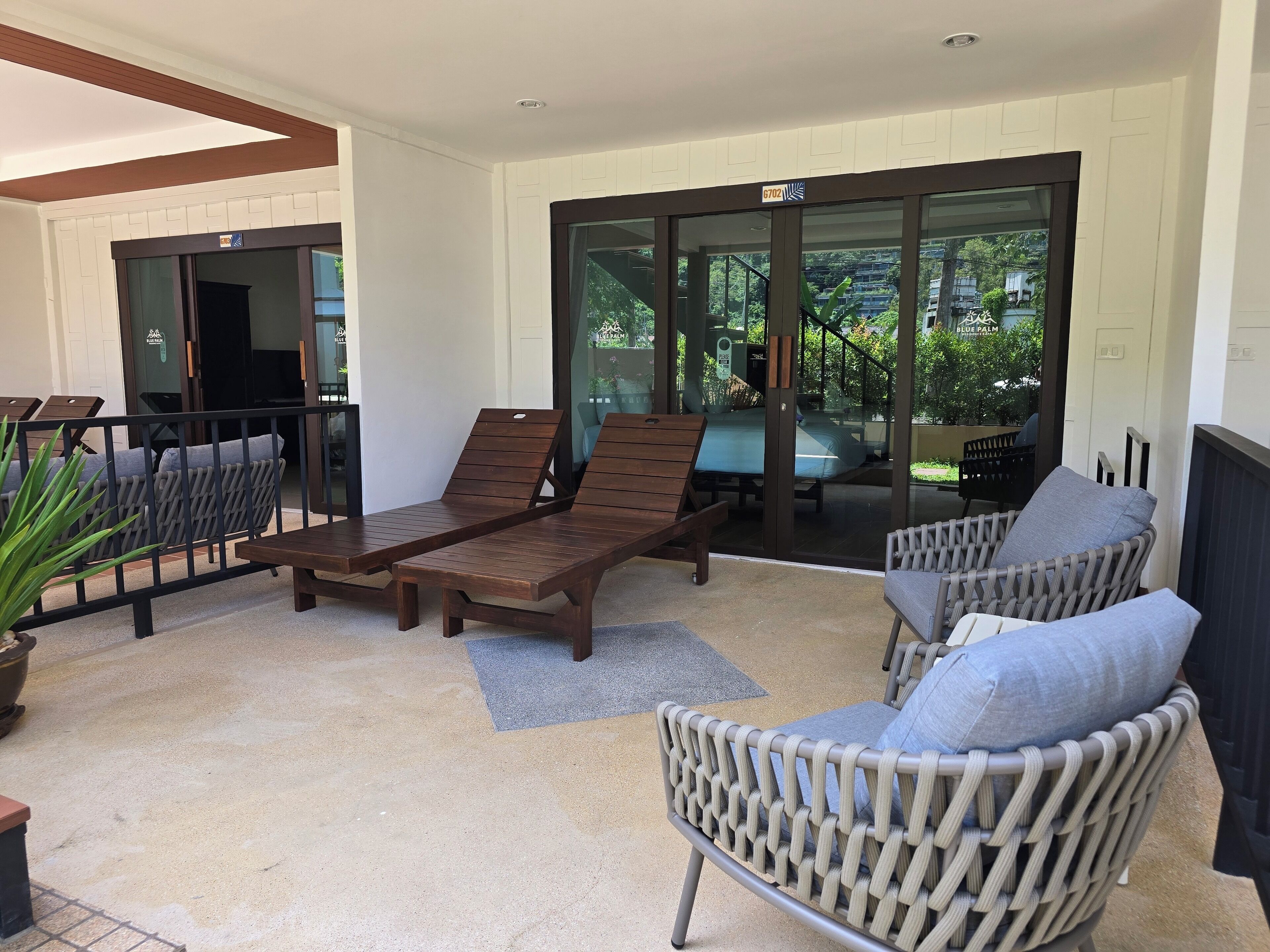 A sunlit outdoor seating area with wooden loungers and wicker chairs, a glass door leading to a pool, and a view of greenery.