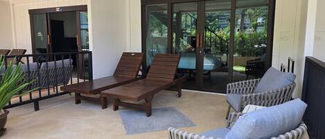 A sunlit outdoor seating area with wooden loungers and wicker chairs, a glass door leading to a pool, and a view of greenery.