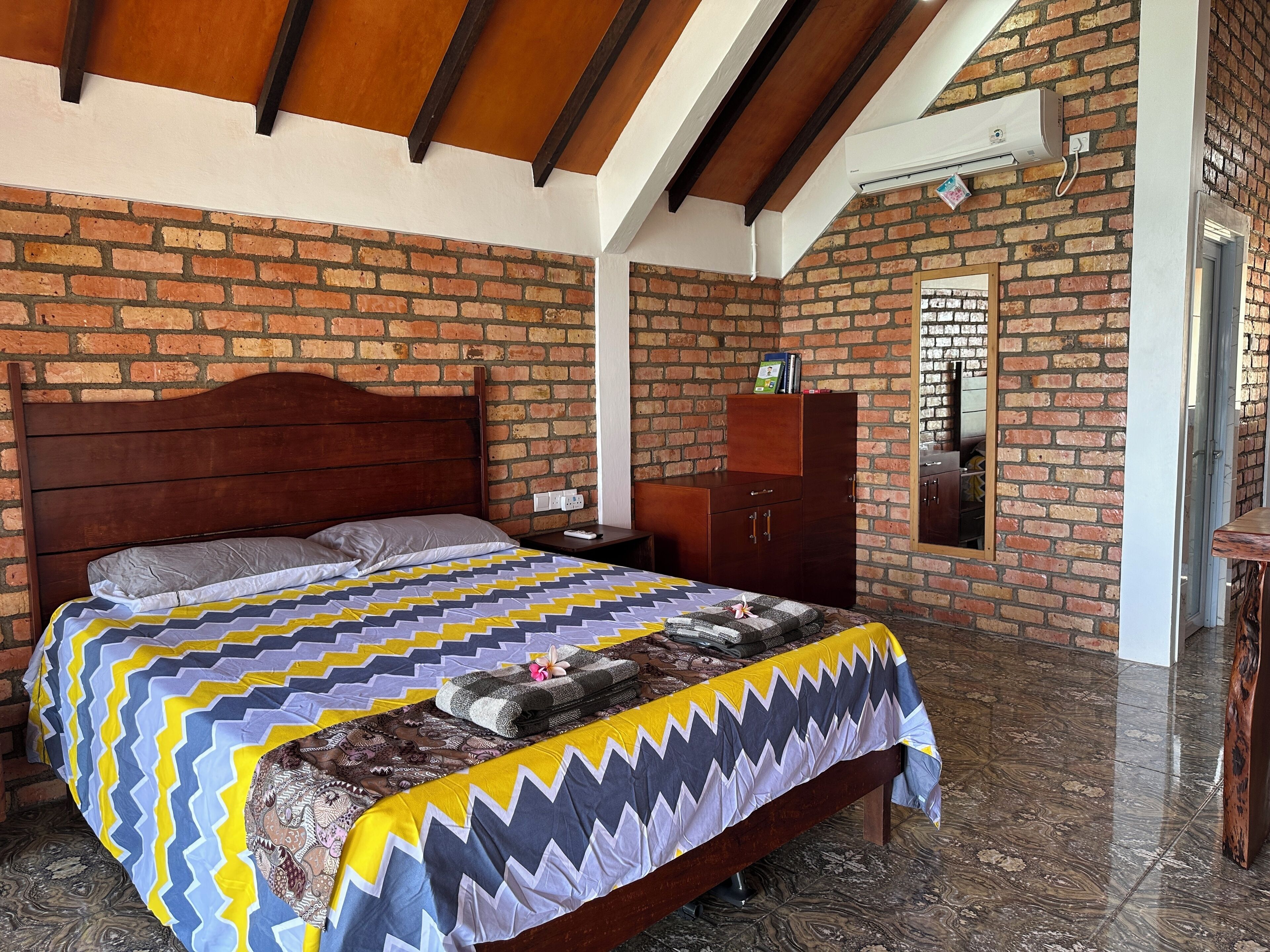 Un dormitorio con pared de ladrillo, vigas de madera en el techo, una cama con colcha estampada, una mesita de noche de madera, un armario y un baño visible a través de una puerta abierta.