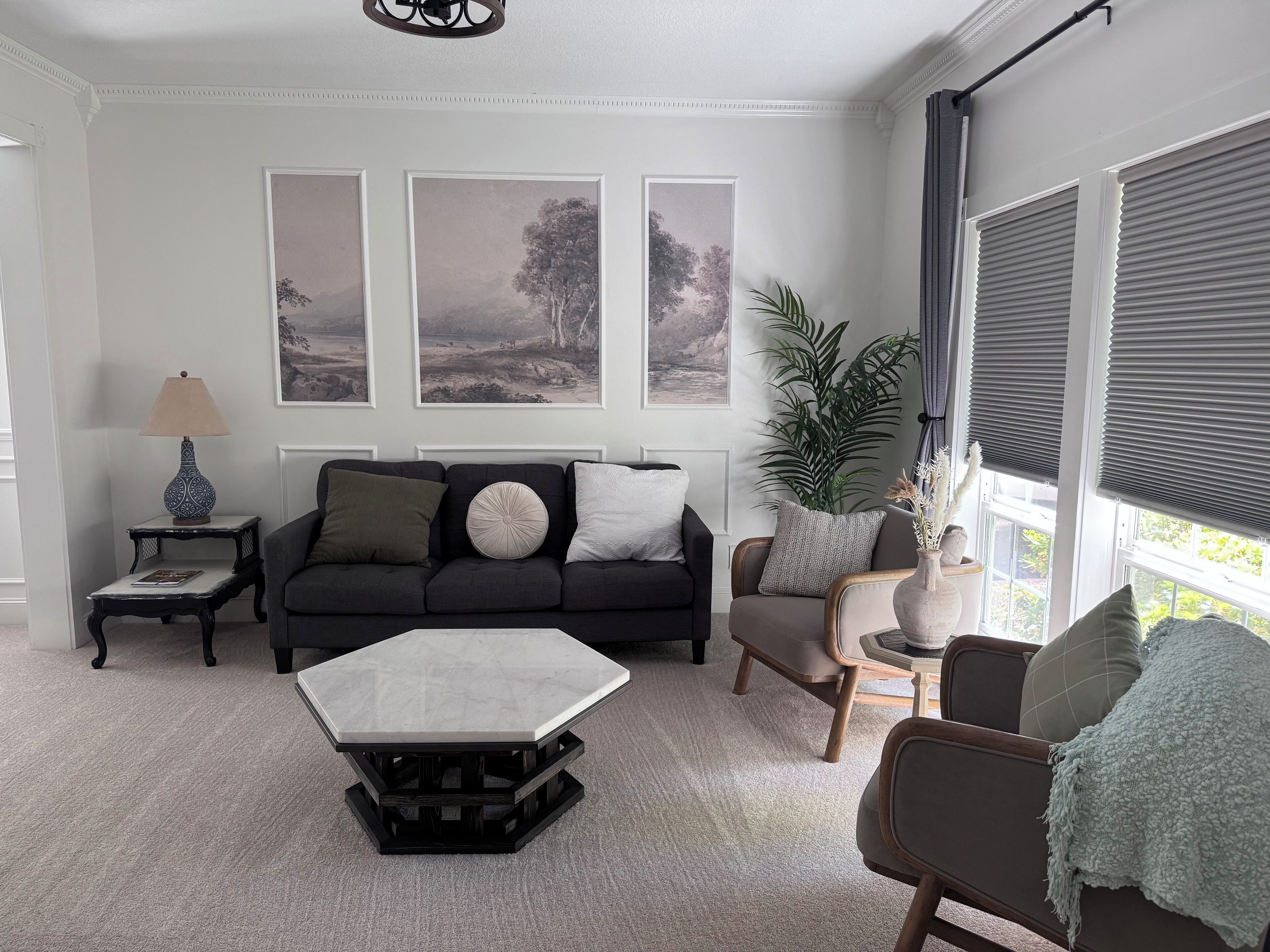Un salon comprenant un canapé, des fauteuils, une table basse et une table d’appoint avec une lampe. Trois tableaux encadrés ornent le mur, et une plante en pot est également présente.