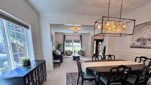 A dining area with a large table, chairs, and a chandelier. There is a potted plant on a console table and a view of the outdoors through a window. - Swim Spa + Hot Tub, Gazebo, BBQ Grill, 6 Bedrooms (Beaverton)