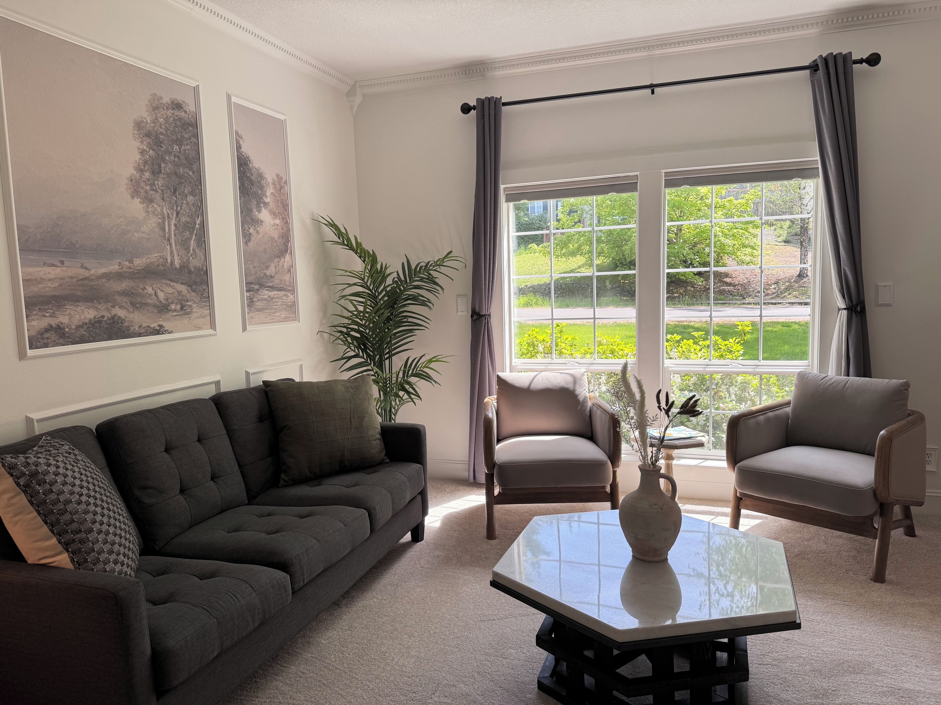A living room with a dark grey sofa, two armchairs, a glass-top coffee table, a potted plant, and a large window with curtains.