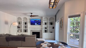 A living room with a fireplace, a television mounted above it, and a dining area with a table set for four. - Swim Spa + Hot Tub, Gazebo, BBQ Grill, 6 Bedrooms (Beaverton)