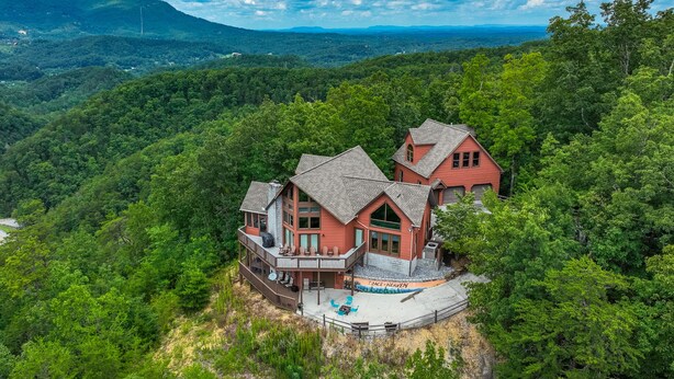 Una casa grande de dos pisos con un porche que rodea toda la estructura y una terraza, rodeada de un bosque frondoso y montañas al fondo.