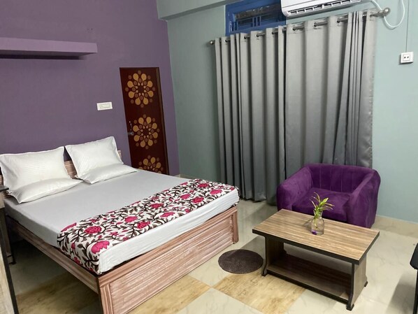 A room with a bed, a purple chair, a small table with a vase, and a window with curtains.