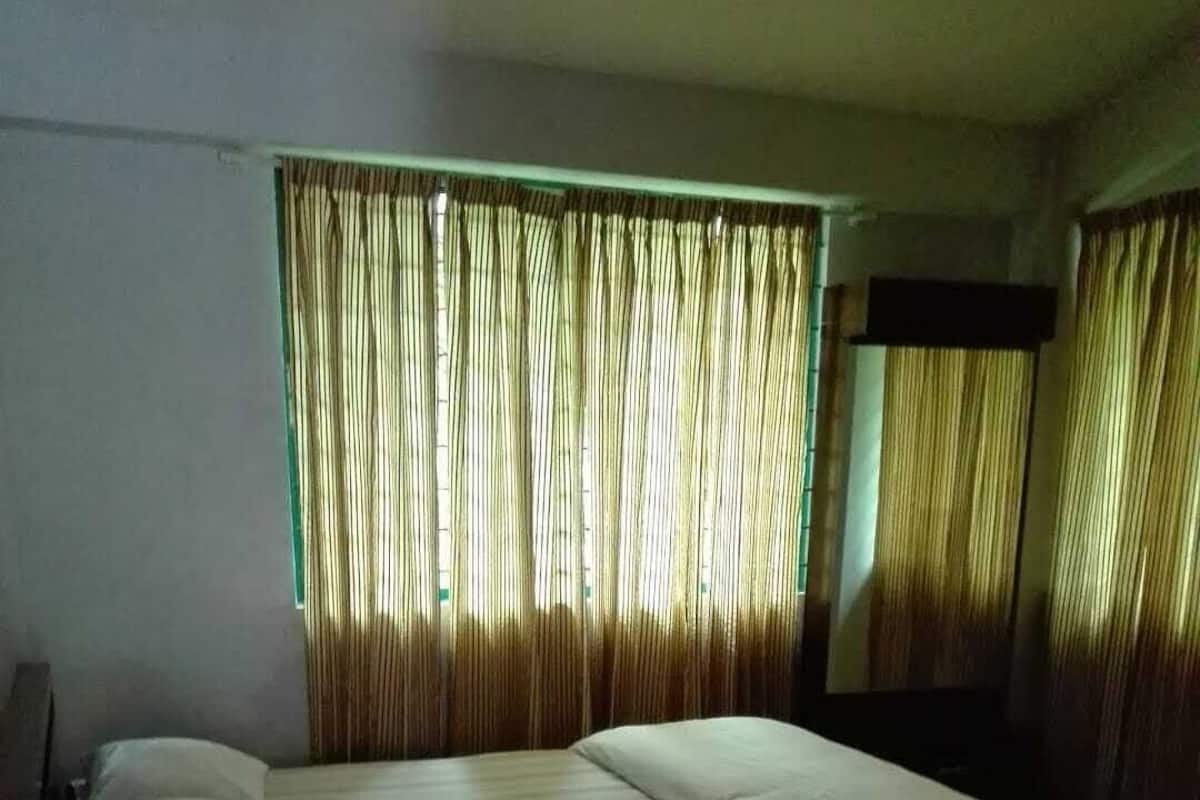 Ein Einzelzimmer mit einem hölzernen Kopfende, einem Nachttisch und einem Fenster mit vertikalen Jalousien.