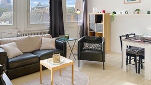 A modern living room with a black leather sofa, a small wooden table, and a black armchair with a patterned cushion. A white radiator is visible, and there is a window with a view of buildings outside.