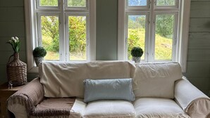 Een gezellige kamer met een witte bank, een klein tafeltje met een vaas en een boek, en twee ramen met uitzicht op groen.