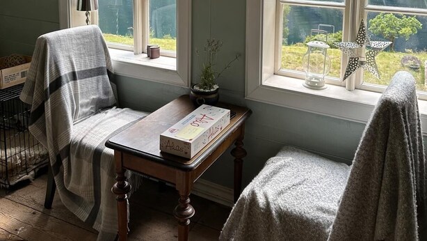 Een kamer met een groot raam, een tafel met een boek en een stoel met een deken.