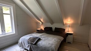 Een slaapkamer met een schuine kap, een groot bed, twee nachtkastjes met lampen, een raam en een vachtkleed.