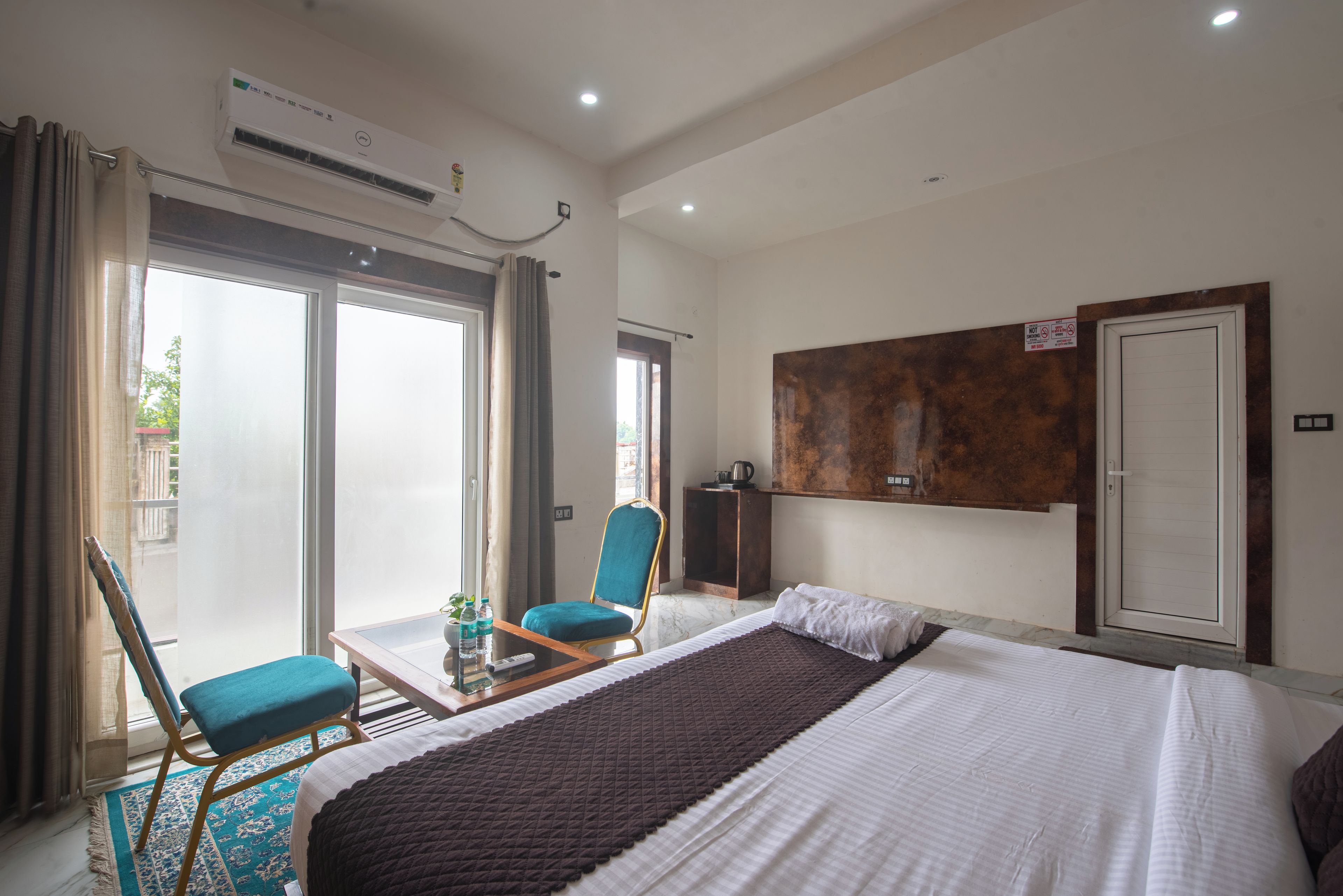 A hotel room with a bed, a chair, a table, an air conditioning unit, and a view of the outside.