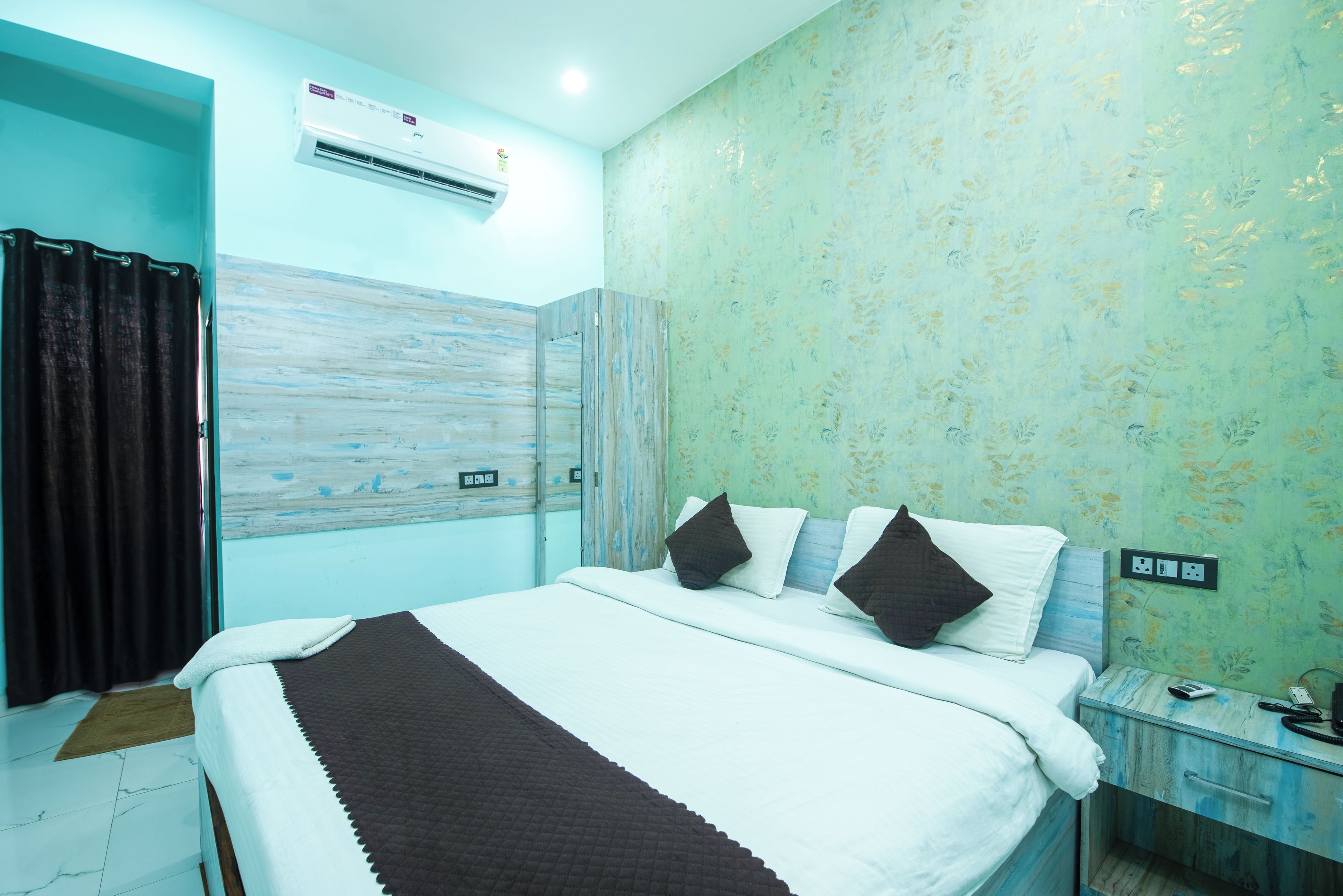 A hotel room with a bed, a bedside table, a nightstand, an air conditioning unit, and a wall with a floral pattern.