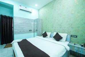 Une chambre d’hôtel comprenant un lit, une table de chevet, une commode, un appareil de climatisation et un mur orné d’un motif floral.