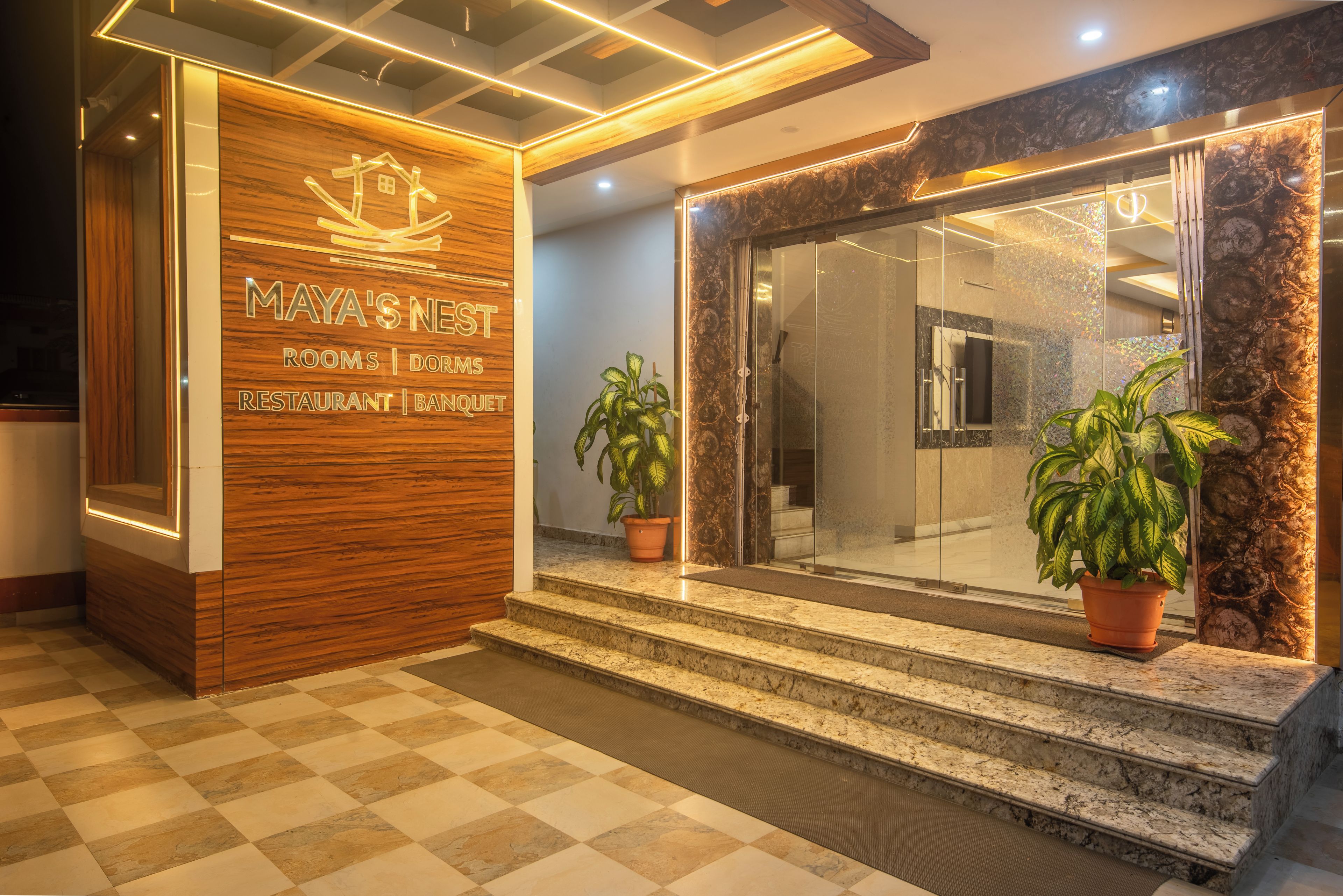 A hotel entrance with a wooden reception desk, a glass door, and potted plants.