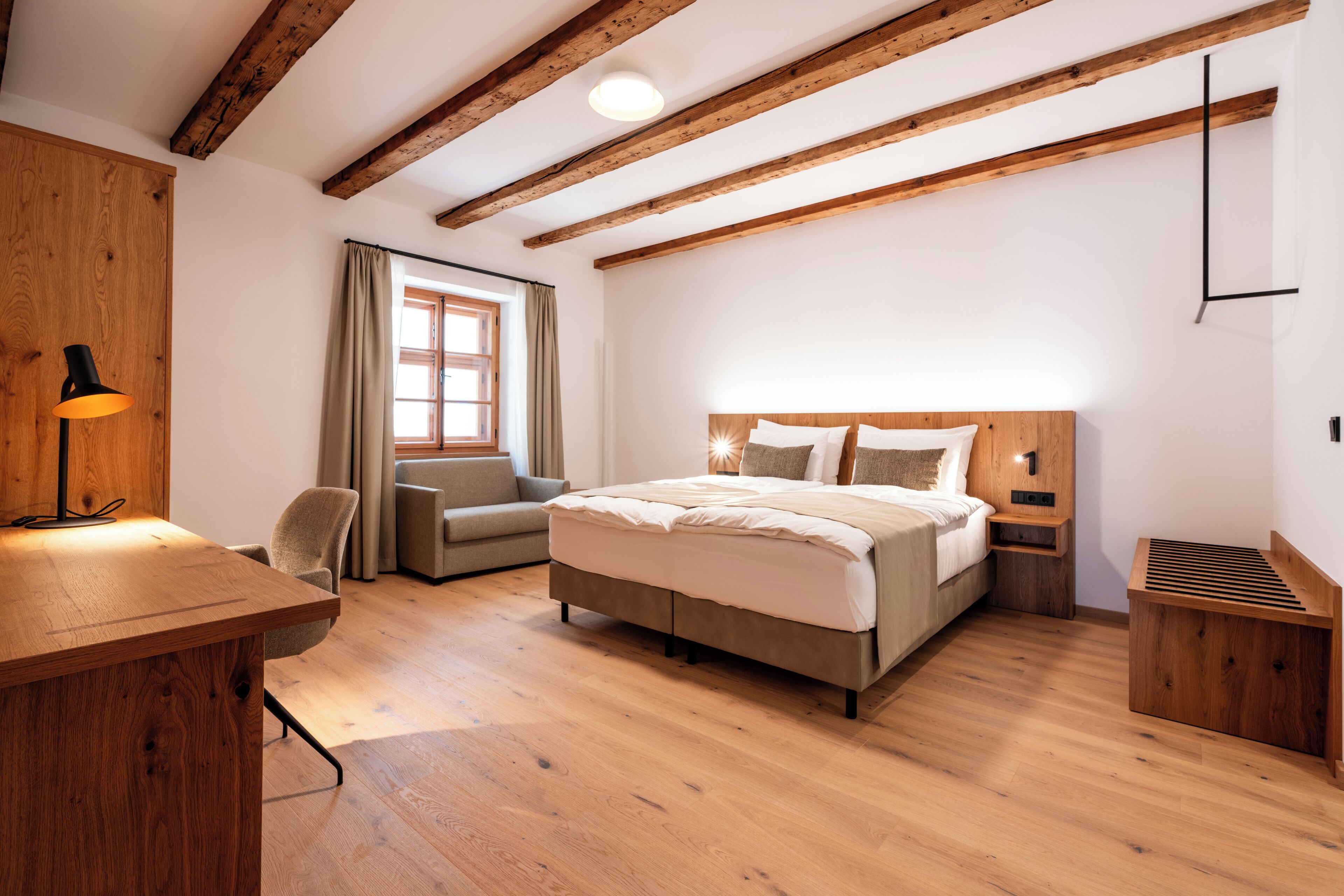 Quarto com teto de madeira, cama com cabeceira, escrivaninha com luminária, uma cadeira e janela com cortinas.