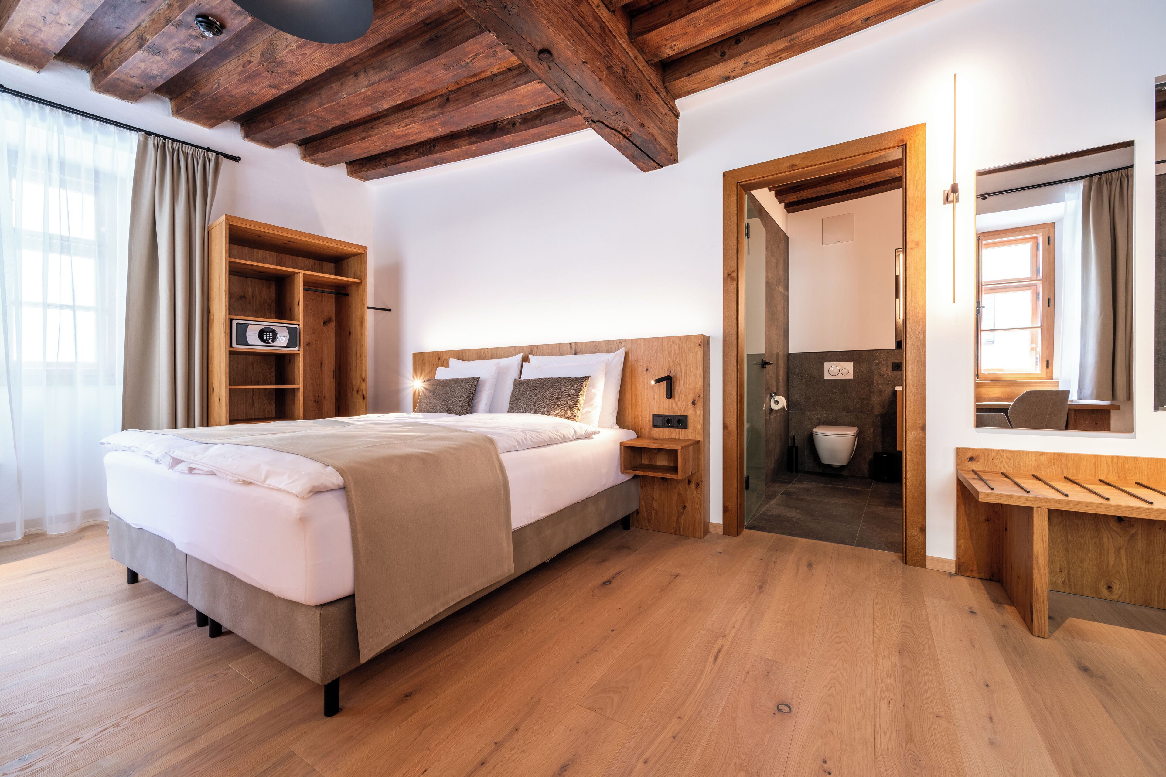 Un dormitorio con cama y mesita de noche de madera, armario de madera, baño con inodoro y lavamanos, y escritorio de madera con espejo.